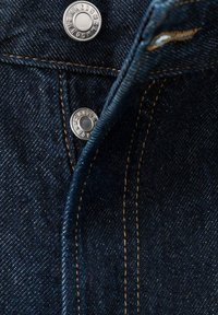 Tela vaquera con dos botones plateados con marca y costuras visibles en naranja en una bragueta de vaqueros parcialmente abierta.