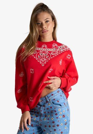 Giovane donna che indossa una felpa rossa con motivi e jeans blu con forme ricamate colorate, posando su uno sfondo semplice.