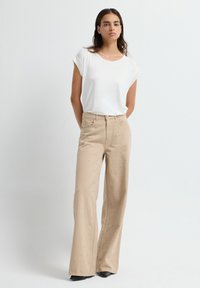 Vrouw die tegen een witte achtergrond staat en een witte top met korte mouwen en beige wijde broek met kleine geborduurde stippen draagt.