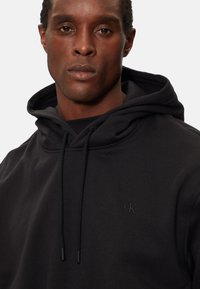 Sudadera negra de tejido suave con una gran capucha, cordones y un sutil logo grabado en el pecho. Diseño simple y moderno.