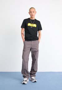 Ung man med tatueringar står med händerna i fickorna, iklädd svart "AIR" Nike t-shirt, grå cargobyxor och vita sneakers på blått golv.