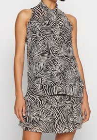Blouse sans manches en imprimé zèbre noir et crème. Présente un col montant avec un nœud et un volant superposé au niveau de l'ourlet. Tissu léger.