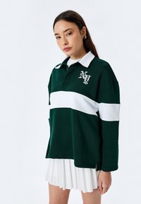 Pullover verde con striscia orizzontale bianca, colletto e testo ricamato. Maniche lunghe, vestibilità rilassata, realizzato in tessuto testurizzato. Gonna plissettata bianca.