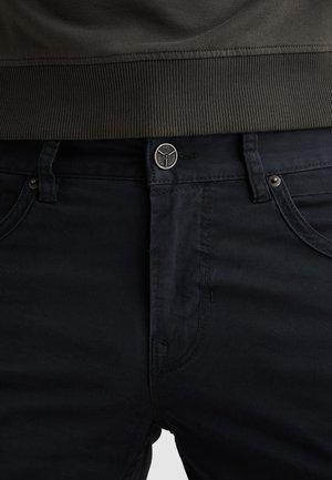 Sezione vita di pantaloni blu scuro con taschino porta monete, chiusura con bottone e un indumento verde oliva scuro a costine infilato nella cintura.