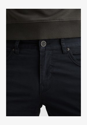 Taillenbereich einer dunkel marineblauen Hose mit Münztasche, Knopfverschluss und einem gerippten dunkel olivgrünen Kleidungsstück, das in den Hosenbund gesteckt ist.