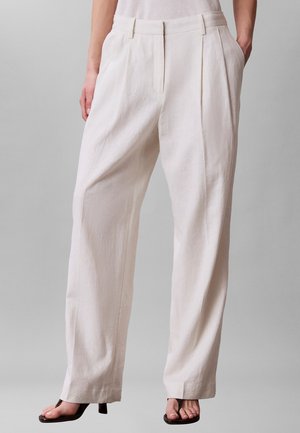 Mujer vestida con pantalones de tiro alto, de corte holgado y color blanco roto, con bolsillos laterales y sandalias negras de tacón y puntera abierta sobre un fondo gris liso.