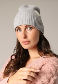 Grijze gebreide beanie met ribbeltextuur en een lichte slouch. Heeft een klein wit label aan de zijkant en is gedragen met een roze trui.