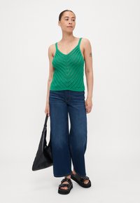 Top de tirantes de punto verde con patrón de V, combinado con unos pantalones vaqueros de pierna ancha de color azul oscuro y sandalias de plataforma negras, junto con un bolso tote negro.