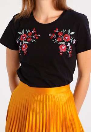 Zwart shirt met korte mouwen en rode en roze bloemrijke borduurwerk op de borst, gecombineerd met een hoog getailleerde geplooide feloranje rok.