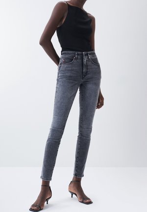 Jean noir délavé slim-fit associé à un top noir sans manches et à des talons ouverts noirs, porté par une personne debout sur un fond uni.