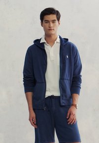 Mornarska zip-up dukserica s prednjim džepom, nošena preko bijele polo majice. U kombinaciji s tamnoplave kratke hlače. Glatka tkanina s ležernim dizajnom.