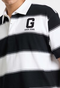 Polo tričko s čiernymi a bielymi prúžkami a bielym golierom, na ľavej hrudi vyšívané písmeno "G" a nápis "GUESS JEANS". Bavlnený materiál.