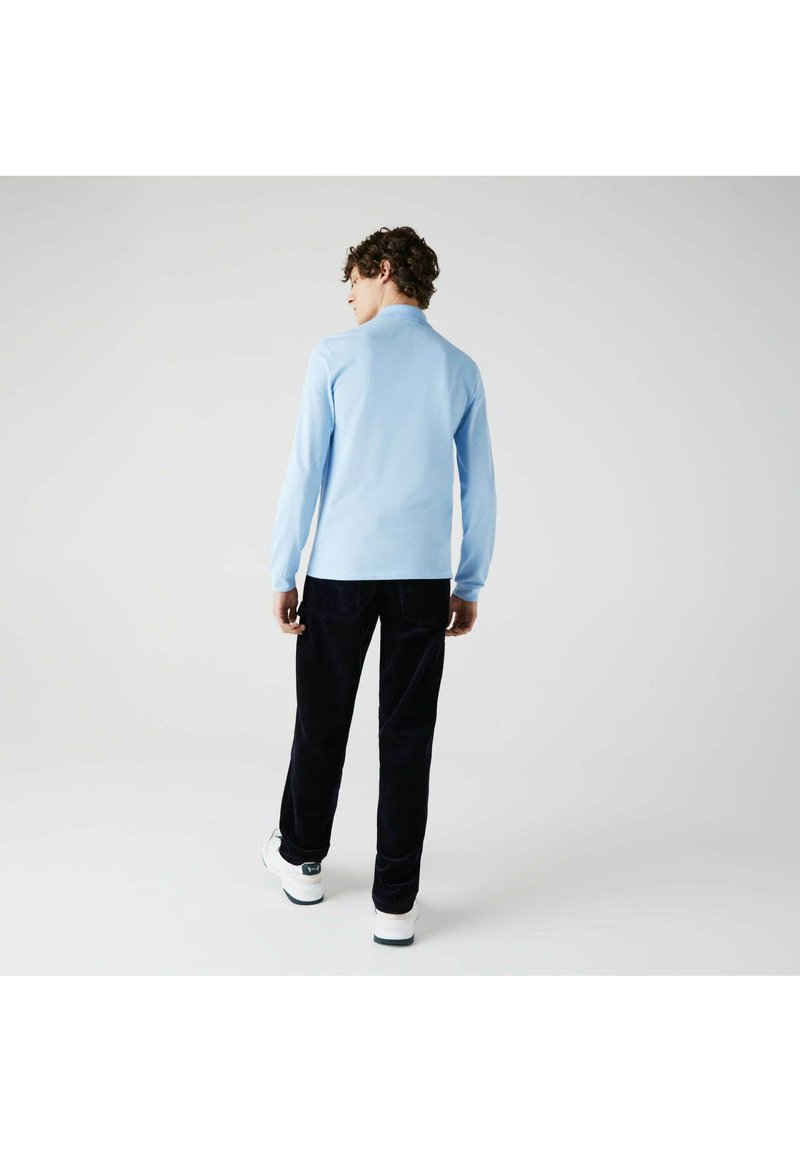 Lysblå langærmet skjorte, med krave, iført sammen med mørke fløjlbukser og hvide sneakers, set fra ryggen mod en neutral baggrund.