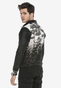 Schwarze Lederjacke mit silbernem gemustertem Rücken, der detaillierte Blumenmuster und metallische Nieten entlang der Naht aufweist.