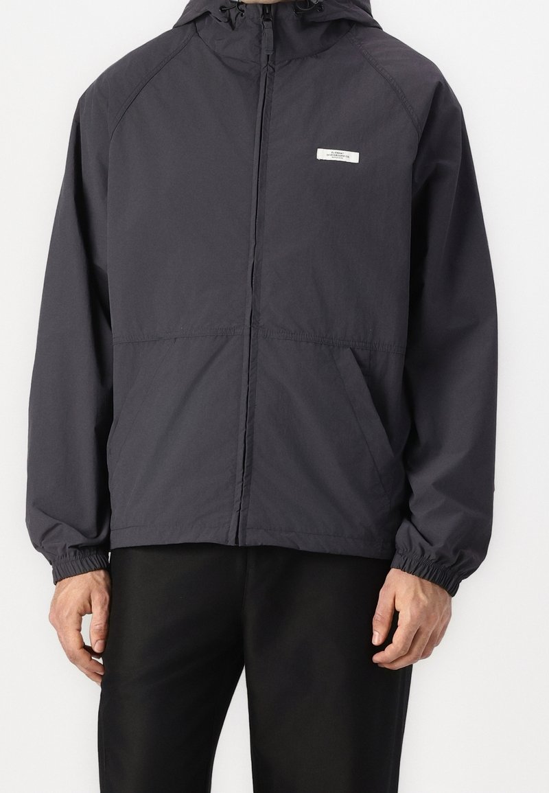 Homme portant une veste noire à capuche avec fermeture éclair, poignets élastiques et poches avant, debout devant un fond blanc.