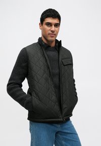 Czarna pikowana kamizelka z wysokim kołnierzem i zamkiem z przodu, noszona na ciemnym swetrze o fakturze, w połączeniu z jasnoniebieskimi dżinsami.