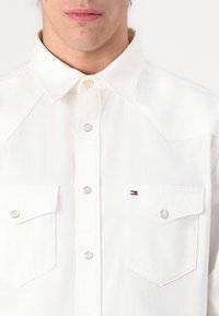 Hvit denimskjorte med trykknapper, spiss krage, to brystlommer og en liten logoakcent i rødt, hvitt og blått.