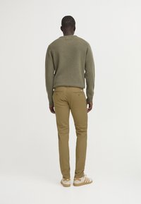 Suéter de punto verde oliva, pantalones de algodón color beige con bolsillos traseros, y zapatos beige con rayas blancas, visto desde atrás.