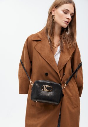 Model in een bruine suede jas, gouden sieraden en een zwarte schoudertas met een logo. Ze kijkt omlaag tegen een witte achtergrond.