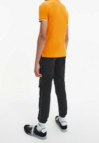 Oranžové polo tričko s bílým lemem, spojené s černými cargo kalhotami s bočními kapsami a bílým prošíváním. Černé tenisky s bílými akcenty.