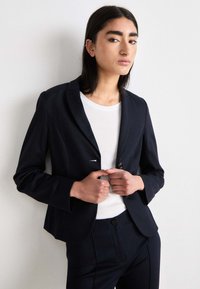 Marineblazer med spiss lapel, enkel knappelukking og sidelommer. Båret over en hvit t-skjorte; kombinert med matchende marinebukser.