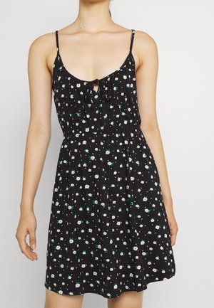 Femme portant une robe noire sans manches avec fines bretelles et petit imprimé floral blanc et vert, debout devant un fond uni clair.