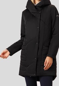 Černý prošívaný středně dlouhý zimní kabát s vysokým límcem, patentkami a přiléhavými rukávy, který nosí osoba s uvolněnýma rukama.