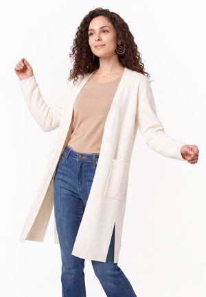 Vrouw met krullend haar draagt een beige top, een lange witte cardigan met voorzakken, en blauwe spijkerbroek, poseert met lichtjes geheven armen.