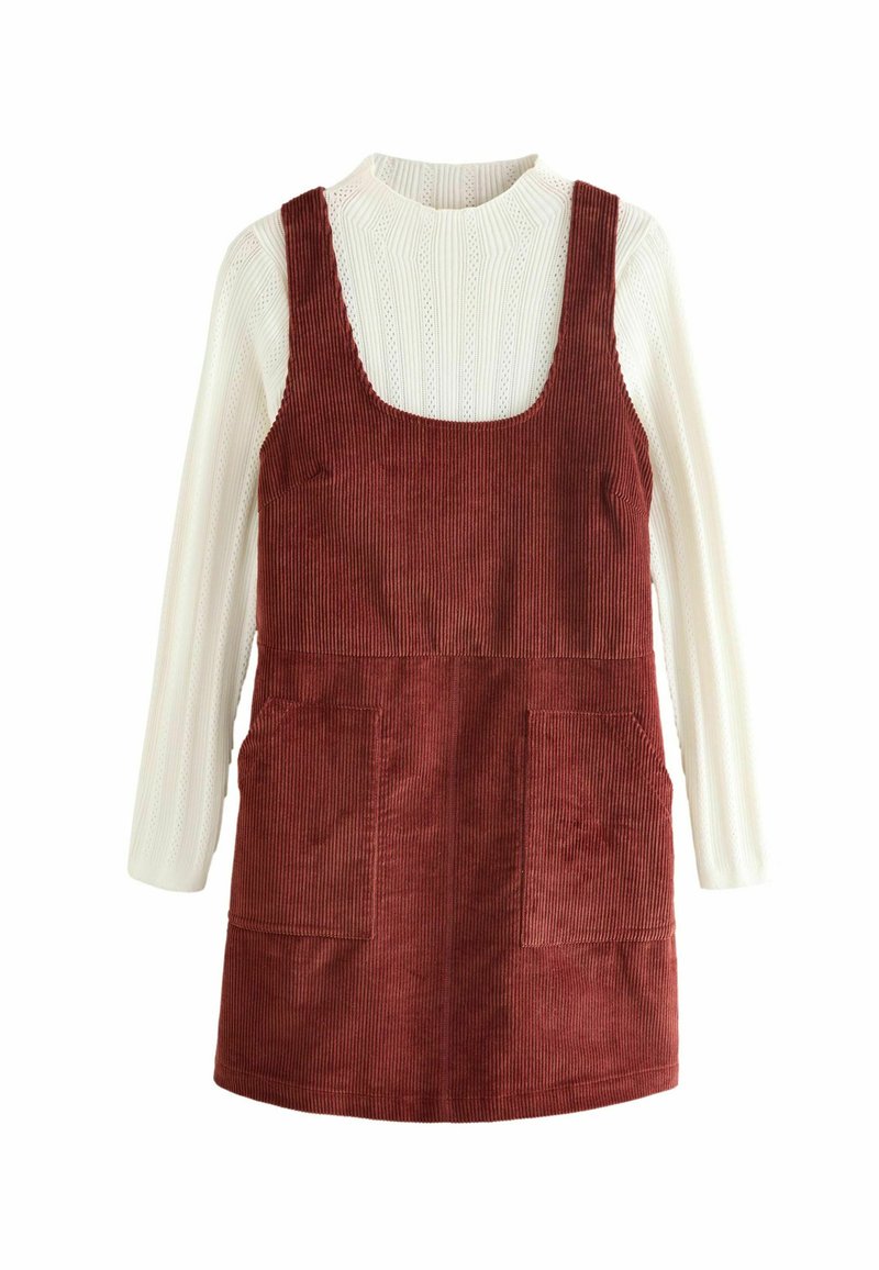 Cord-U-Roy Latzkleid in Rost mit quadratischem Ausschnitt, zwei vorderen Taschen und einem gerippten weißen Langarmoberteil darunter.