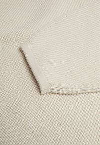 Beige strukturiertes Gewebe mit einem gerippten Muster und einem glatten Besatz. Die Nahaufnahme zeigt eine gefaltete Kante und eine Nähdetail an der Naht.