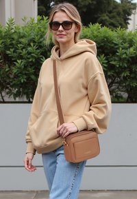 Felpa con cappuccio color sabbia, dotata di una tasca frontale, abbinata a un paio di jeans azzurri chiari. Il modello indossa una piccola borsa a tracolla marrone con una superficie texture.
