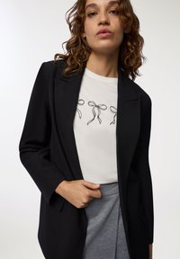 Blazer nero con rever a punta sopra una maglietta bianca con grafiche di nastri neri. Gonna grigia a righe sottili. Texture del tessuto lisce visibili.