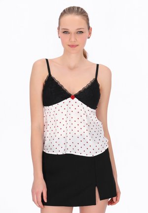 Világos hajú nő hátrafogott hajjal, fekete, csipkével díszített, fehér pöttyös spagetti pántos felsőt és elöl sliccelt fekete szoknyát visel.