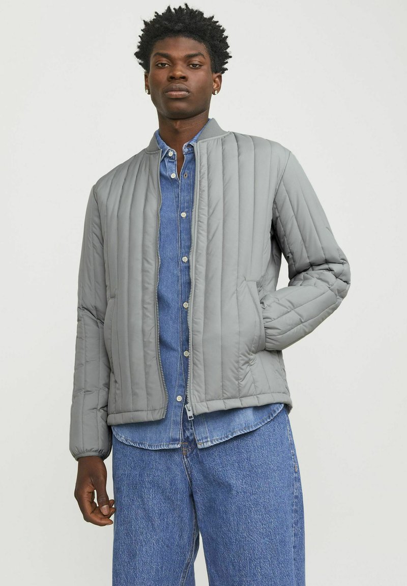 Giacca grigia trapuntata con design bomber, caratterizzata da un colletto a coste e chiusura con zip, indossata sopra una camicia in denim blu e pantaloni.