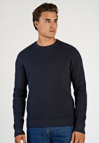 Marineblauer gerippter Strickpullover mit Rundhalsausschnitt, langen Ärmeln und enganliegendem Saum, kombiniert mit blauen Jeans. Glatte Textur.