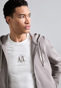 Grå zip-up hoodie med blød tekstur, der har et frontlogo. Underneath en tætsiddende hvid t-shirt med broderet branding i beige.