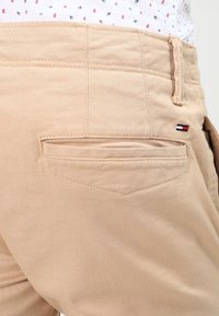 Beige cotton pants with a back pocket, featuring a small branded logo in red, white, and blue. The fabric has a smooth texture.