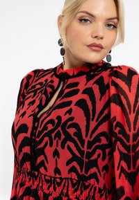 Rotes und schwarzes gemustertes Kleid aus strukturiertem Stoff, mit puffärmel und einem knopfartigen Ausschnitt mit gerafftem Taillendetail.