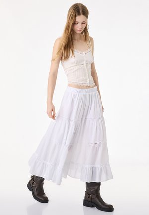 Giovane donna che indossa un top senza maniche bianco con rifiniture in pizzo, gonna lunga bianca a balze e stivaletti scuri, su uno sfondo semplice.