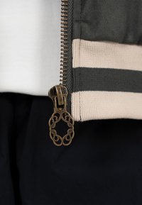 Gros plan d'un curseur de fermeture éclair en métal avec une boucle ornée attachée à un bord de tissu rayé noir et beige sur un vêtement.