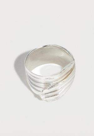 Anillo ancho de plata con diseño texturizado y en capas y detalle asimétrico superpuesto sobre un fondo claro y liso.