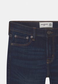 Granatowe dżinsy z rozszerzanym dołem, z żółtymi szwami, dwoma przednimi kieszeniami oraz metką wskazującą rozmiar 9/10.