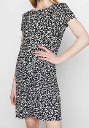Femme portant une robe noire à manches courtes avec un motif floral blanc, posant avec une main sur la hanche sur un fond clair uni.