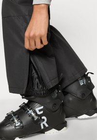 Sorte skibukser med elastiske manchetter, lynlåsedetalje ved kanten, båret over sorte skistøvler med metalliske spænder og hvid tekst.