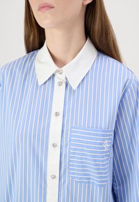 Camicia a righe azzurro chiaro con colletto bianco, chiusura frontale con bottoni decorativi e taschino anteriore con un logo discreto.