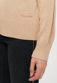 Suéter de punto beige con puños y dobladillo acanalados; presenta una pequeña etiqueta con la inscripción "A|X Armani Exchange". Combinado con pantalones vaqueros negros que exhiben un acento de cremallera.