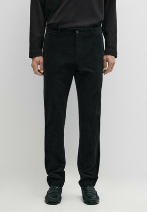 Uomo che indossa pantaloni neri dritti, camicia nera a maniche lunghe e mocassini neri, in piedi su uno sfondo semplice.