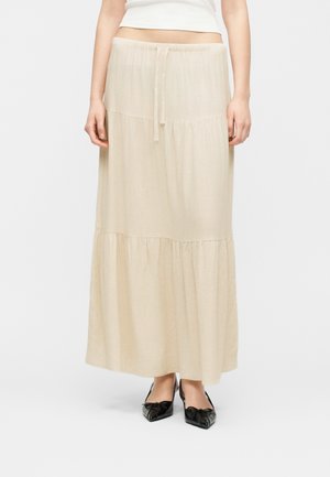 Jupe longue beige à volants avec taille à cordon, portée avec des ballerines noires à bout pointu et un haut court blanc.