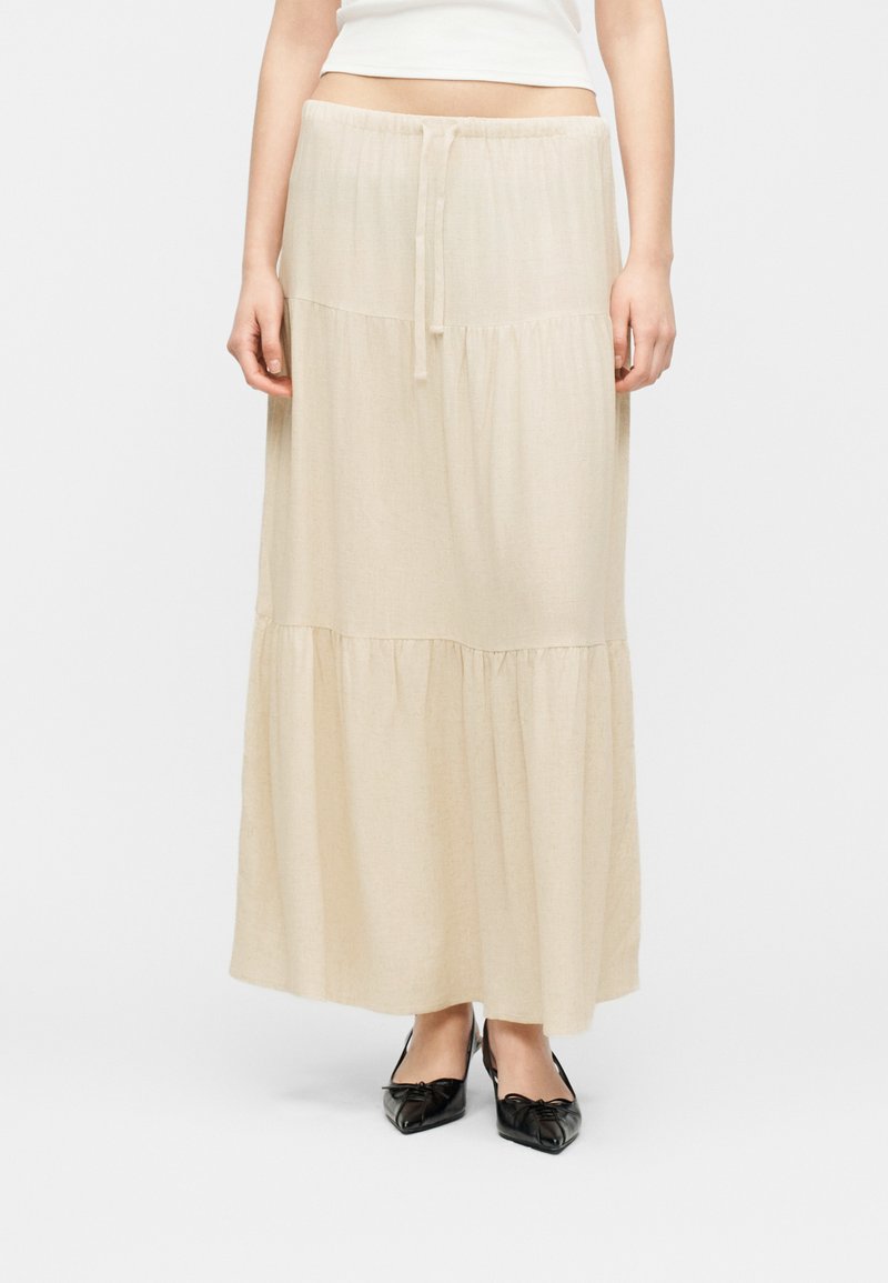 Jupe longue beige à volants avec taille à cordon, portée avec des ballerines noires à bout pointu et un haut court blanc.