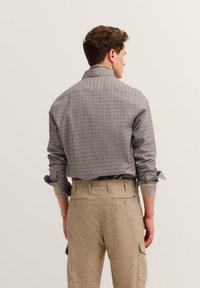 Mann, der mit dem Rücken zur Kamera steht, trägt ein grau kariertes Hemd mit hochgekrempelten Ärmeln und beige Hosen mit Knöpfen an der Rückseite und seitlichen Taschen.